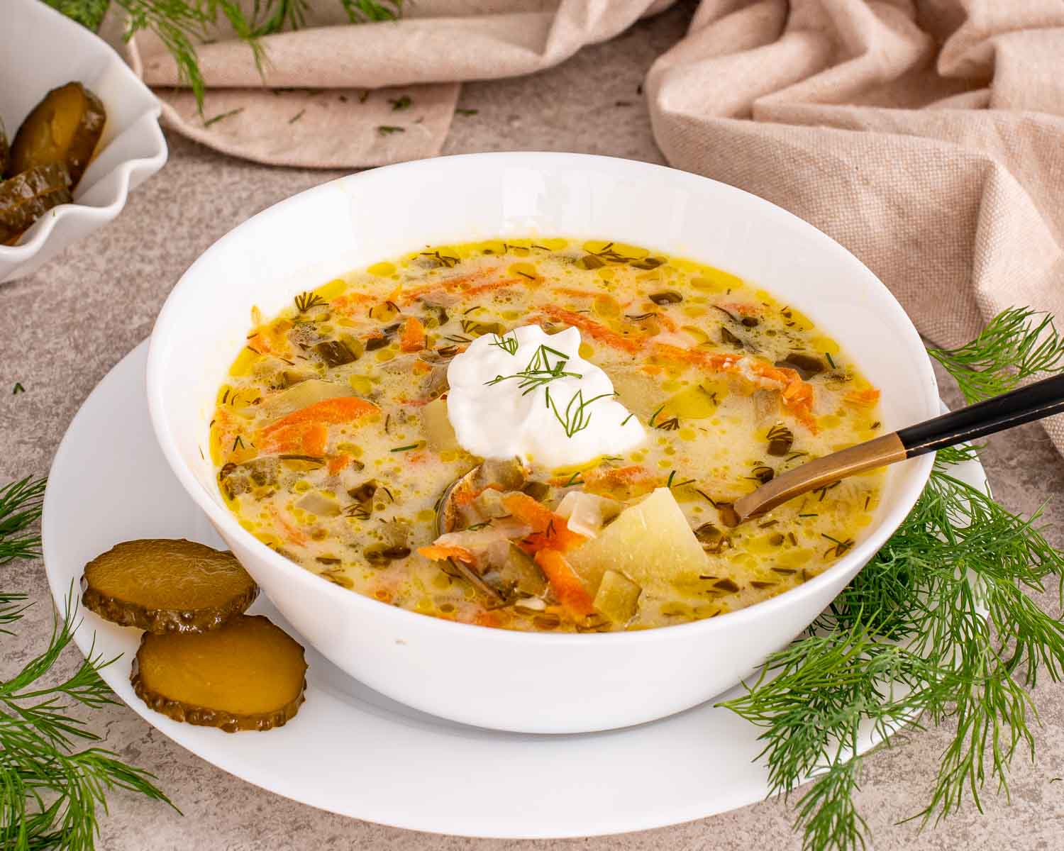 Creamy dill pickle soup in white bowl with sliced pickles, herbs, and spoon showing potatoes and vegetables in broth.