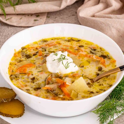 Dill pickle soup served in white bowl with sour cream, potatoes, carrots, chopped pickles, and fresh dill garnish.