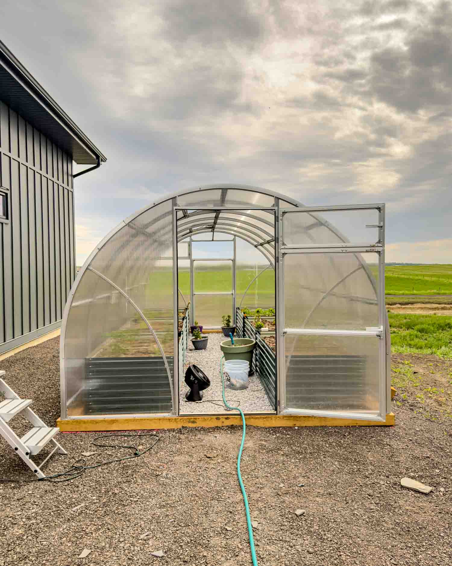 Front view of a polycarbonate greenhouse with both doors open wide revealing the interior raised beds and Alberta prairie fields stretching to the horizon beyond.