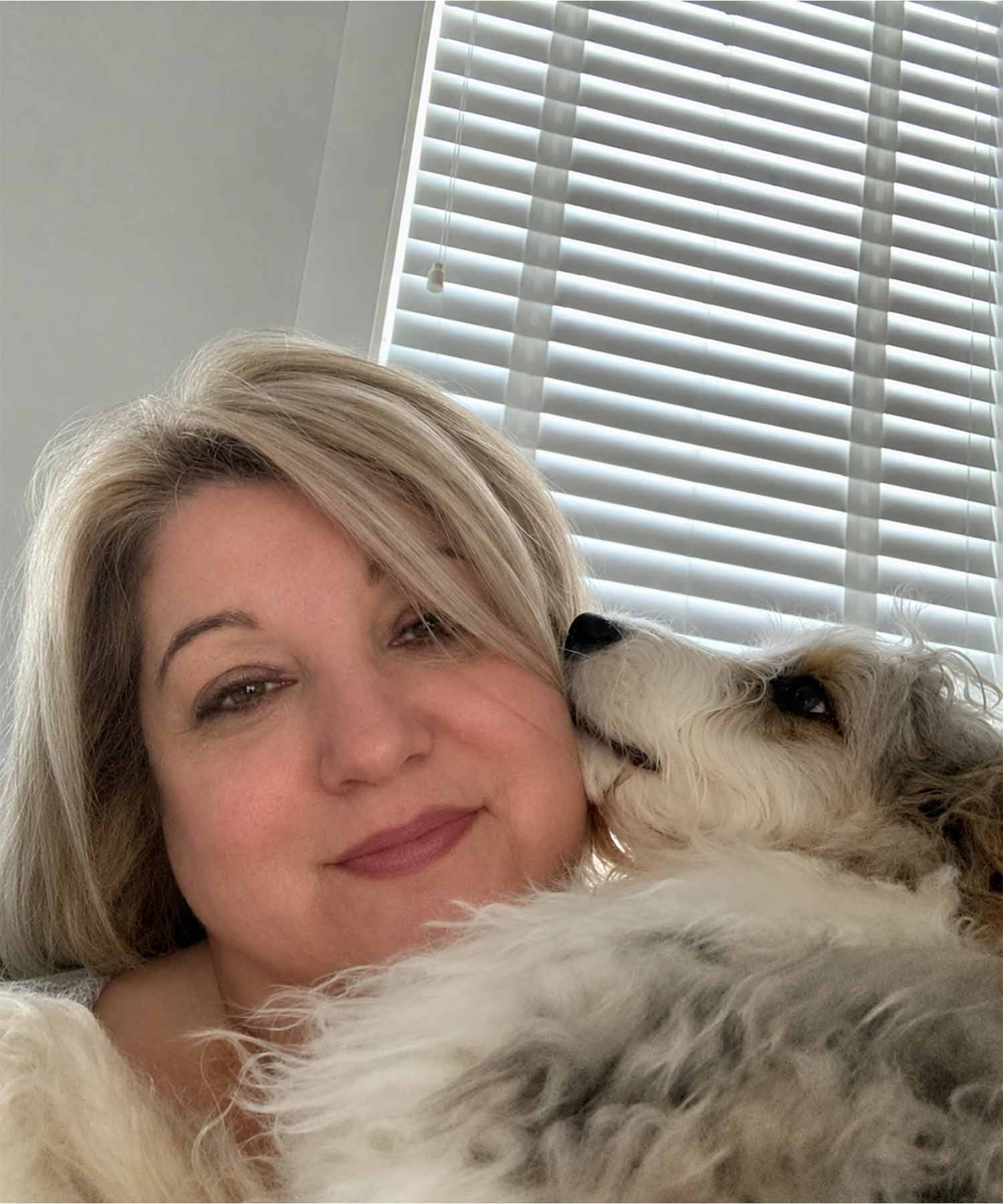 Close up selfie of Jo smiling with a fluffy grey and white dog nuzzling against her cheek by a window with white blinds.