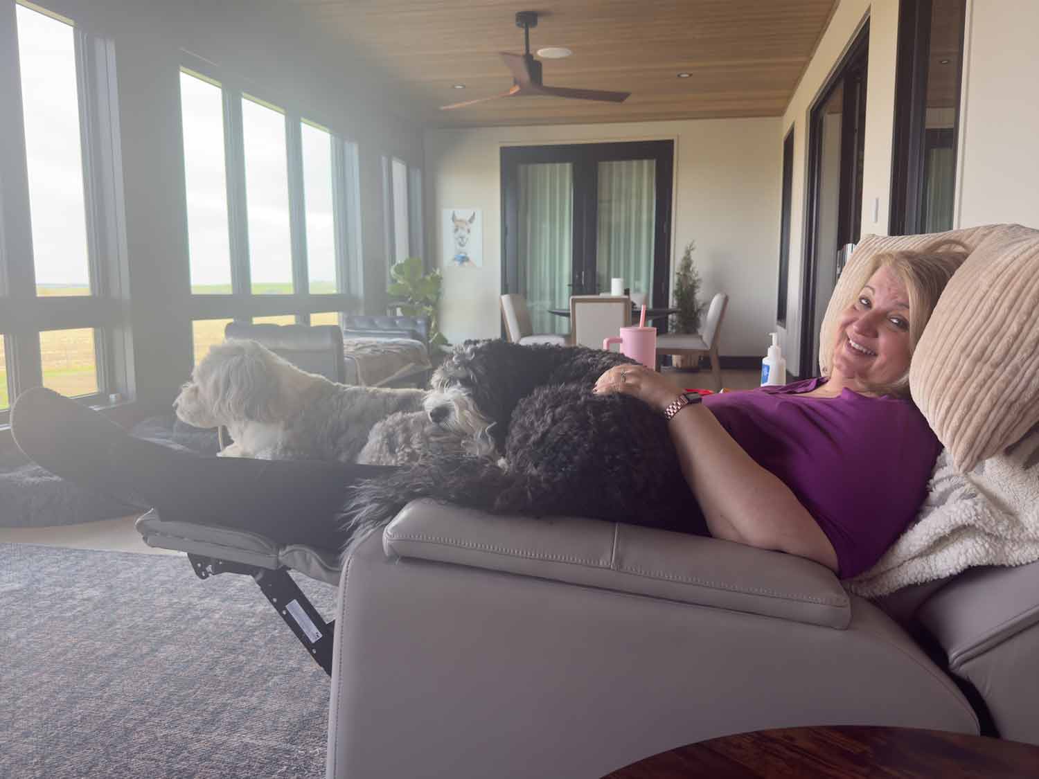 Jo reclining in the sunroom with two large fluffy dogs draped across her legs in a bright room with floor to ceiling windows overlooking fields.