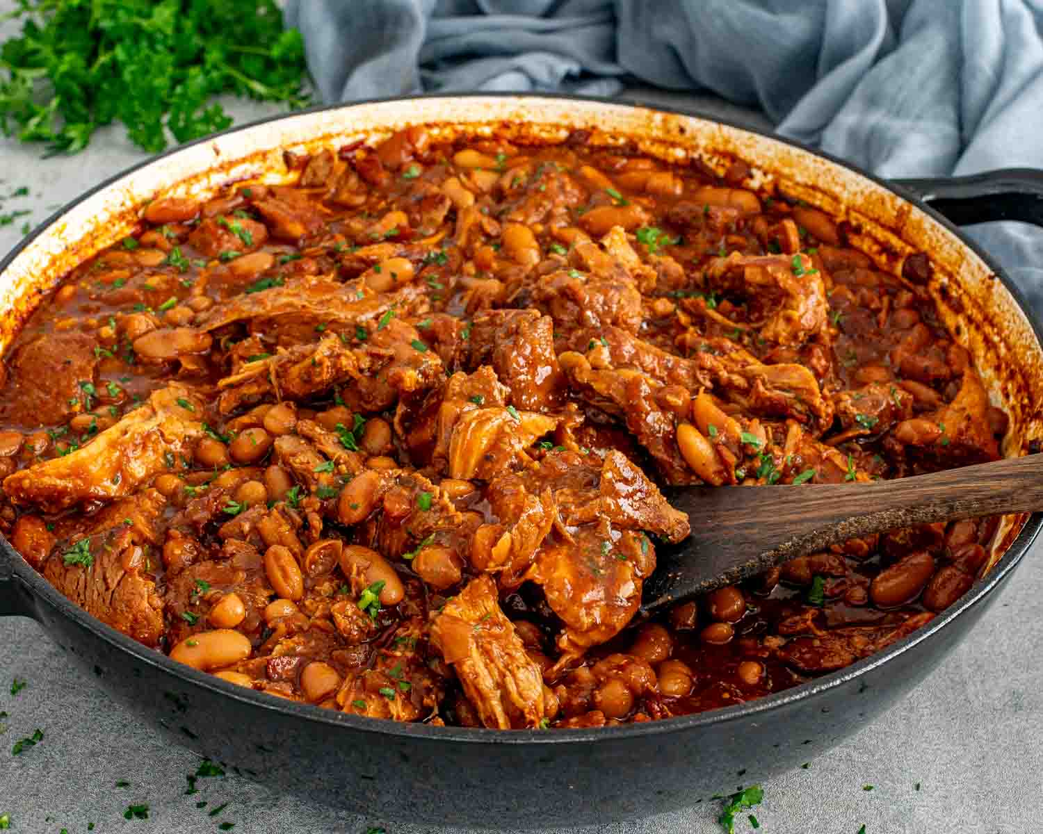 Homemade pork and beans with pulled pork and navy beans in a rich smoky sauce in a dark braising pan with a wooden spoon and fresh parsley.