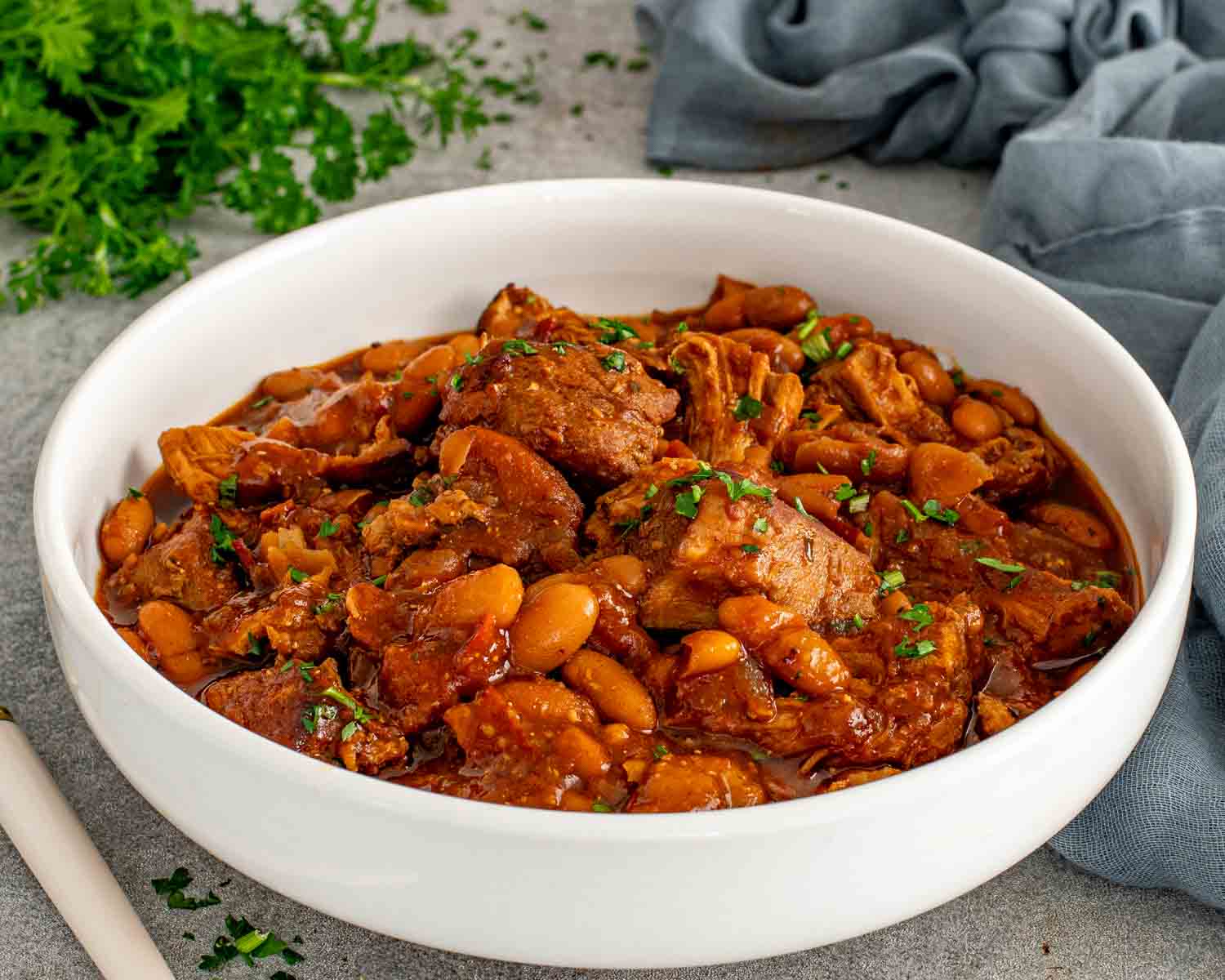 Homemade pork and beans with chunks of tender pork shoulder and navy beans in a smoky sauce served in a white bowl garnished with chopped fresh parsley.