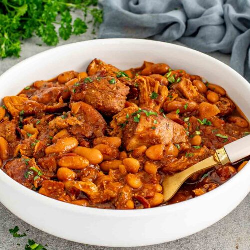 Homemade pork and beans with tender pulled pork shoulder and navy beans in a rich smoky sauce served in a white bowl with a gold spoon and fresh parsley.