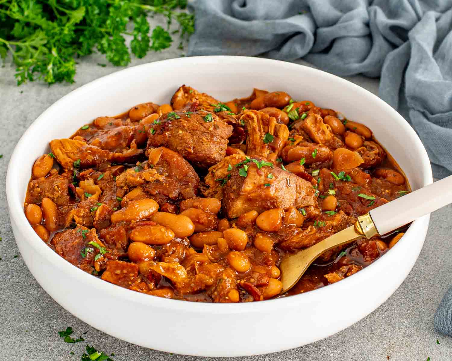 Homemade pork and beans with tender pulled pork shoulder and navy beans in a rich smoky sauce served in a white bowl with a gold spoon and fresh parsley.