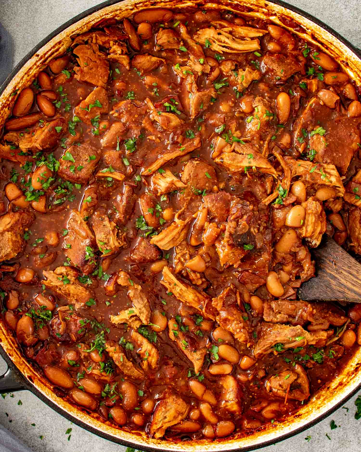 Overhead view of homemade pork and beans with pulled pork and navy beans in a rich smoky sauce in a Dutch oven with a wooden spoon.