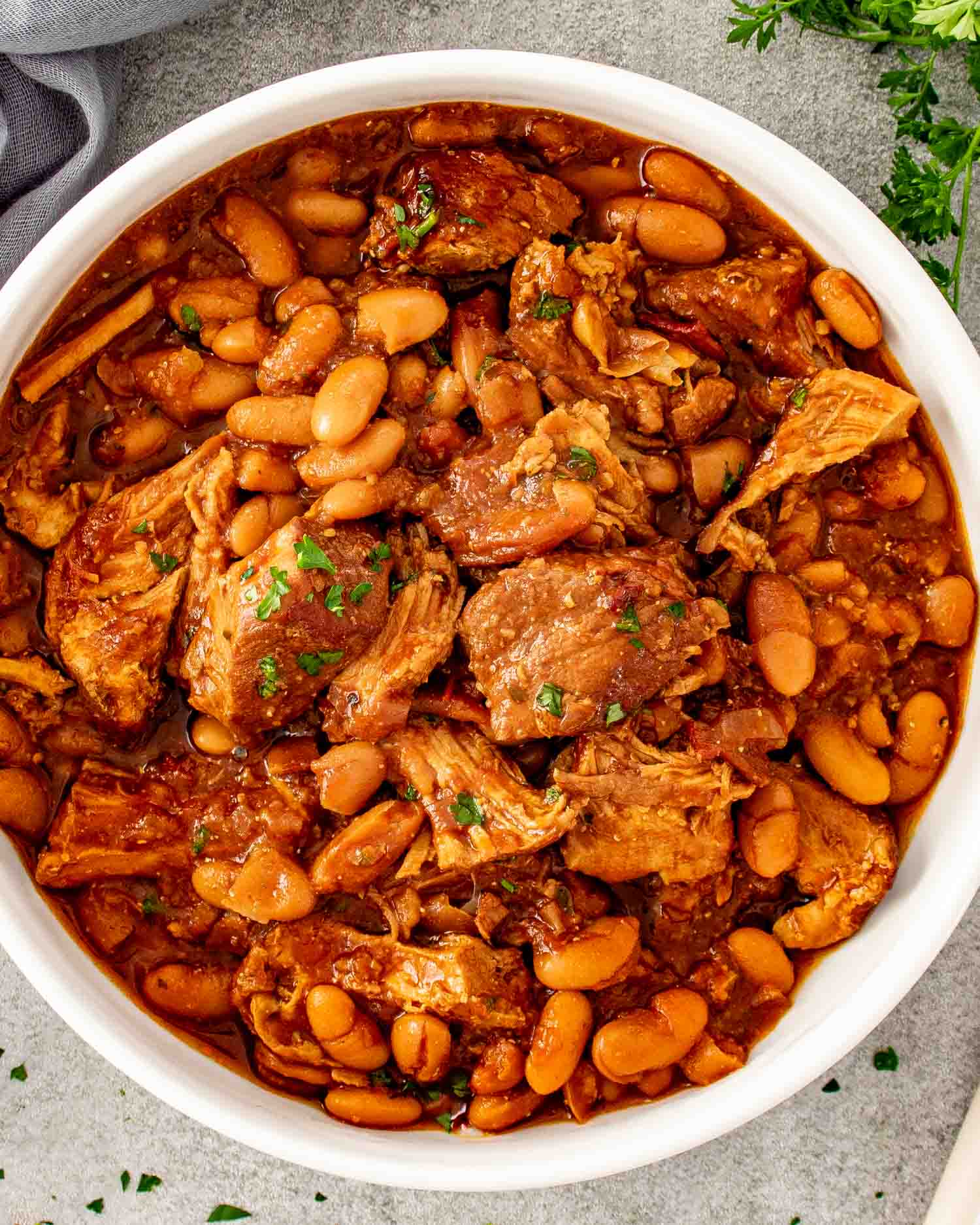 Overhead view of homemade pork and beans with pulled pork shoulder and navy beans in a smoky sauce served in a large white bowl garnished with fresh parsley.