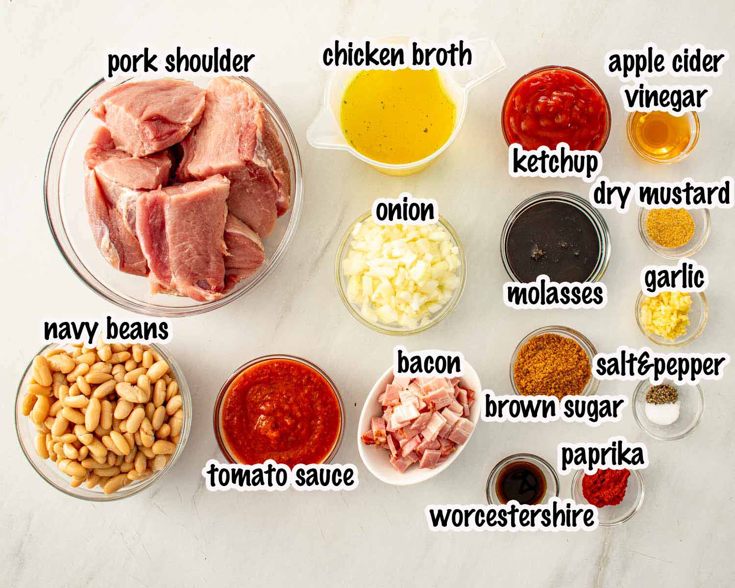 All ingredients for homemade pork and beans laid out on a white surface including pork shoulder, navy beans, bacon, tomato sauce, molasses, chicken broth, onion, garlic, ketchup, and spices.