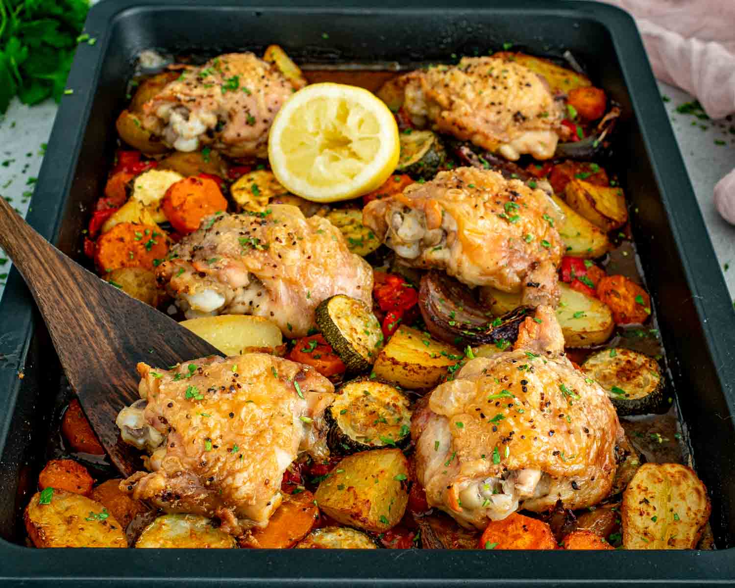Six bone in chicken thighs roasted over baby potatoes, carrots, zucchini, and red onion on a dark sheet pan with a halved lemon and wooden spatula.