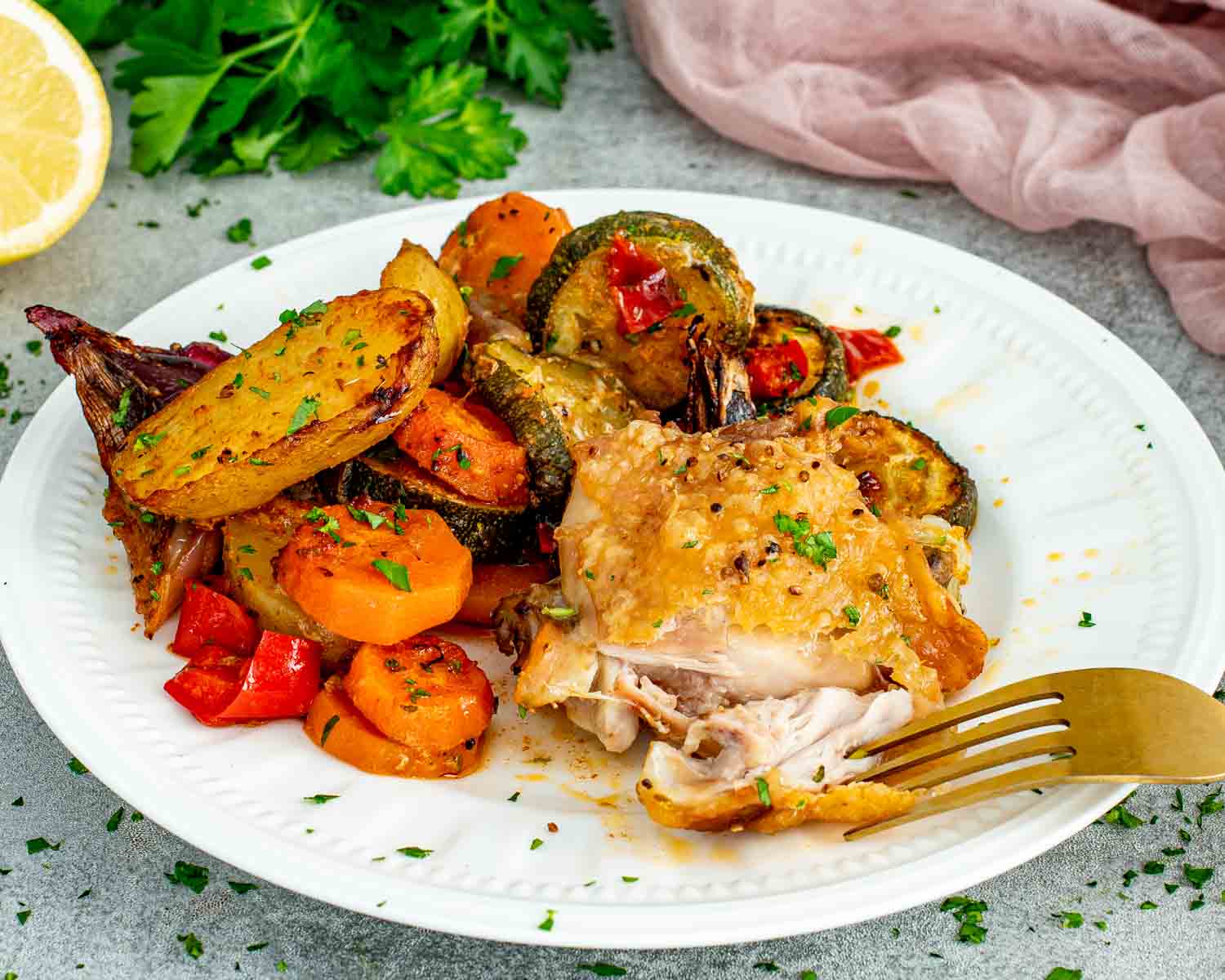 A roasted chicken thigh pulled apart with a gold fork on a white plate alongside caramelized baby potatoes, zucchini, carrots, and red bell pepper with fresh parsley and lemon in the background.