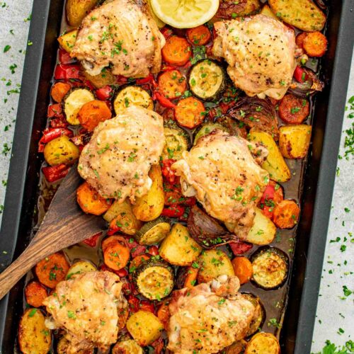 Overhead view of 6 roasted chicken thighs on a full sheet pan with baby potatoes, carrots, zucchini, red bell pepper, red onion, a halved lemon, and fresh parsley.
