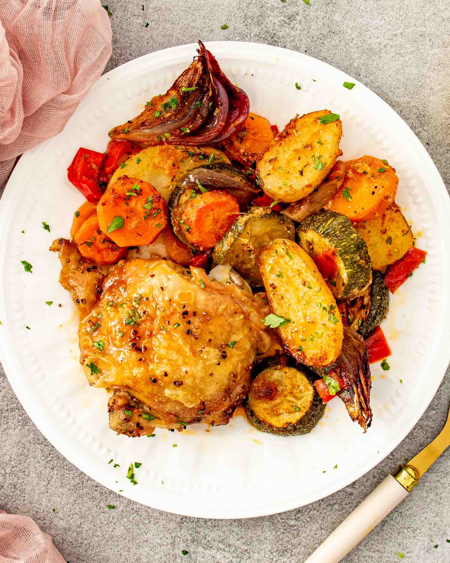 Overhead view of a golden roasted chicken thigh on a white plate with caramelized baby potatoes, zucchini, carrots, red bell pepper, and red onion garnished with fresh parsley.
