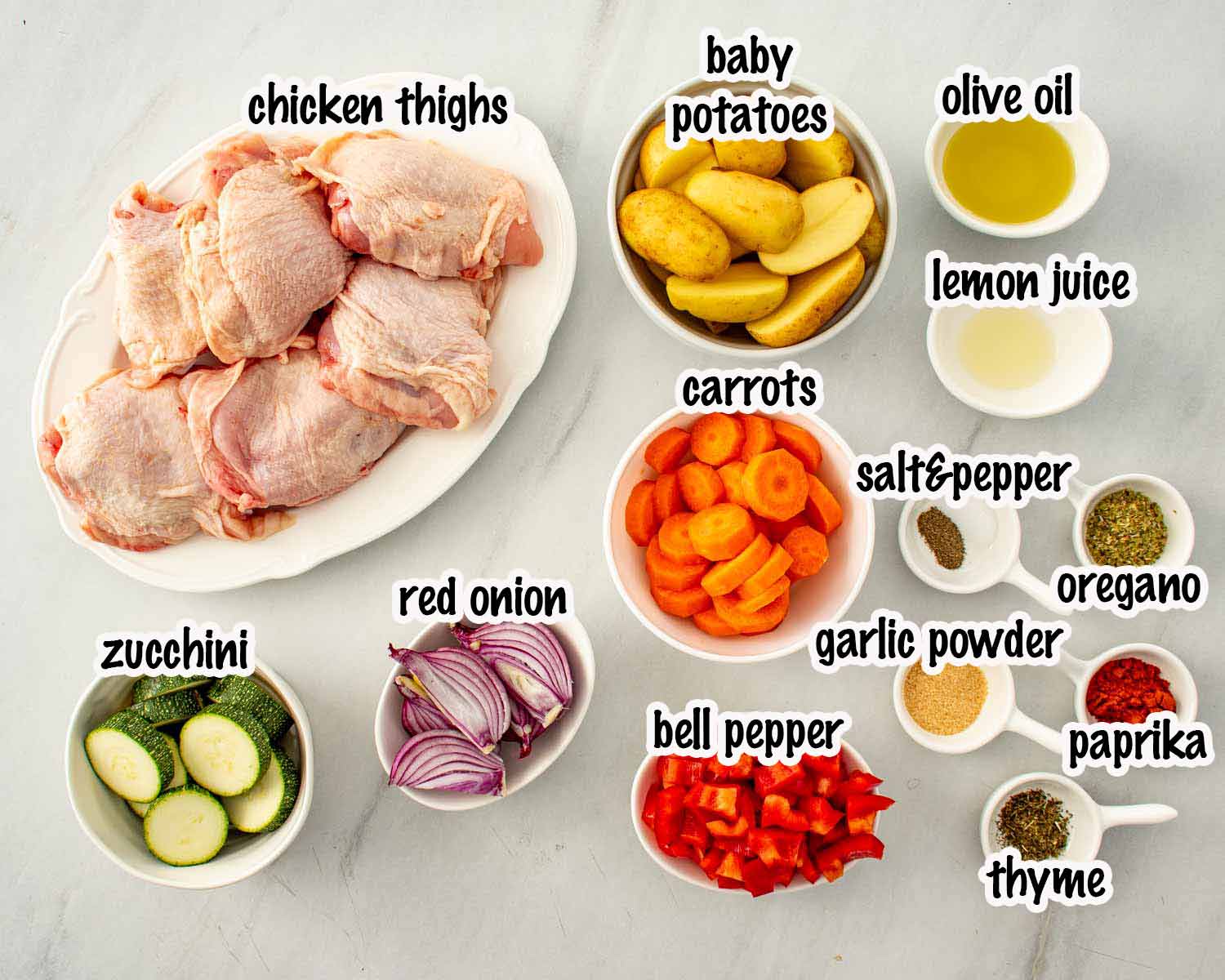 Overhead flat lay of sheet pan chicken thigh ingredients including bone in chicken thighs, baby potatoes, carrots, zucchini, red onion, red bell pepper, olive oil, lemon juice, paprika, garlic powder, oregano, thyme, salt, and pepper.