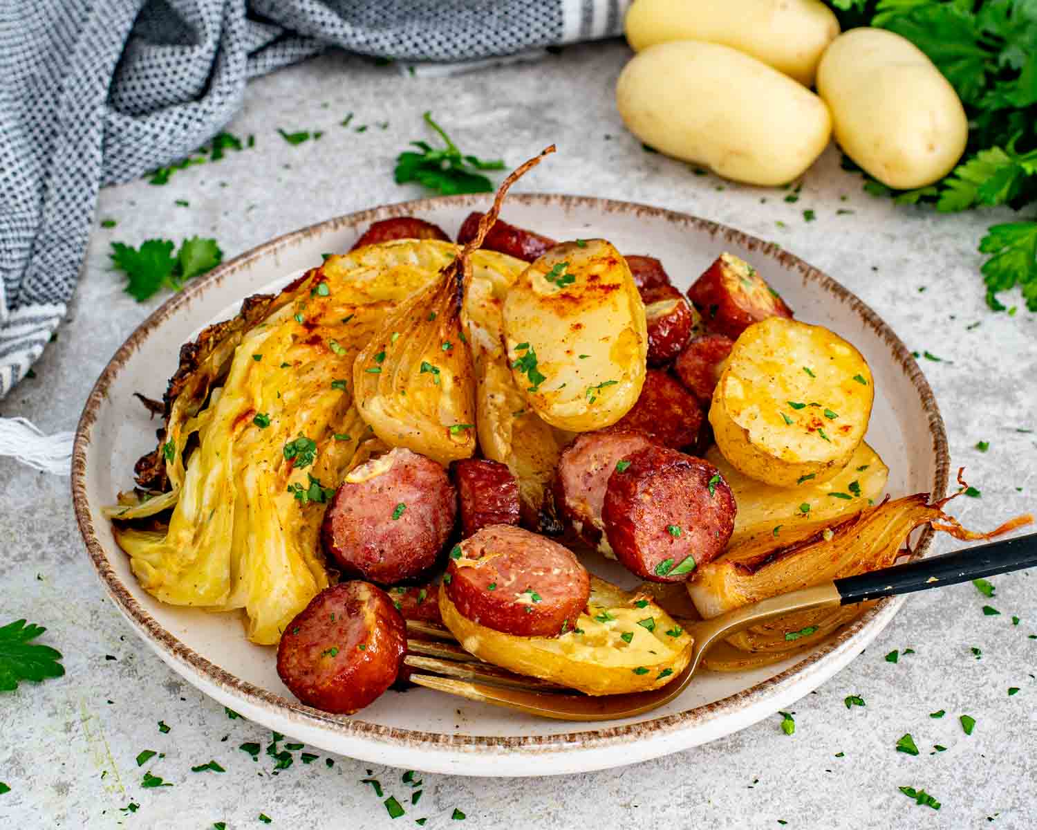 A single serving of sheet pan sausage and cabbage with roasted baby potatoes, caramelized onion, and a gold fork on a rustic ceramic plate.