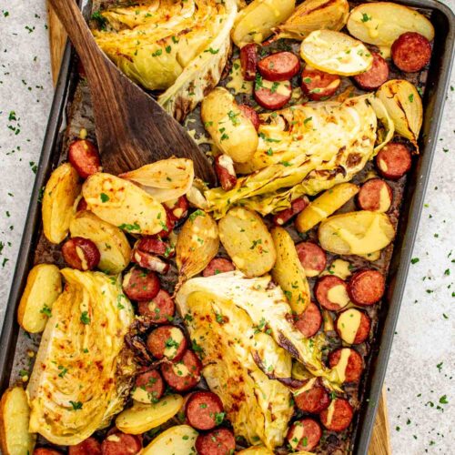 Overhead sheet pan of roasted sausage, caramelized cabbage wedges, baby potatoes, and onion with Dijon apple cider drizzle and fresh parsley on a wood board.