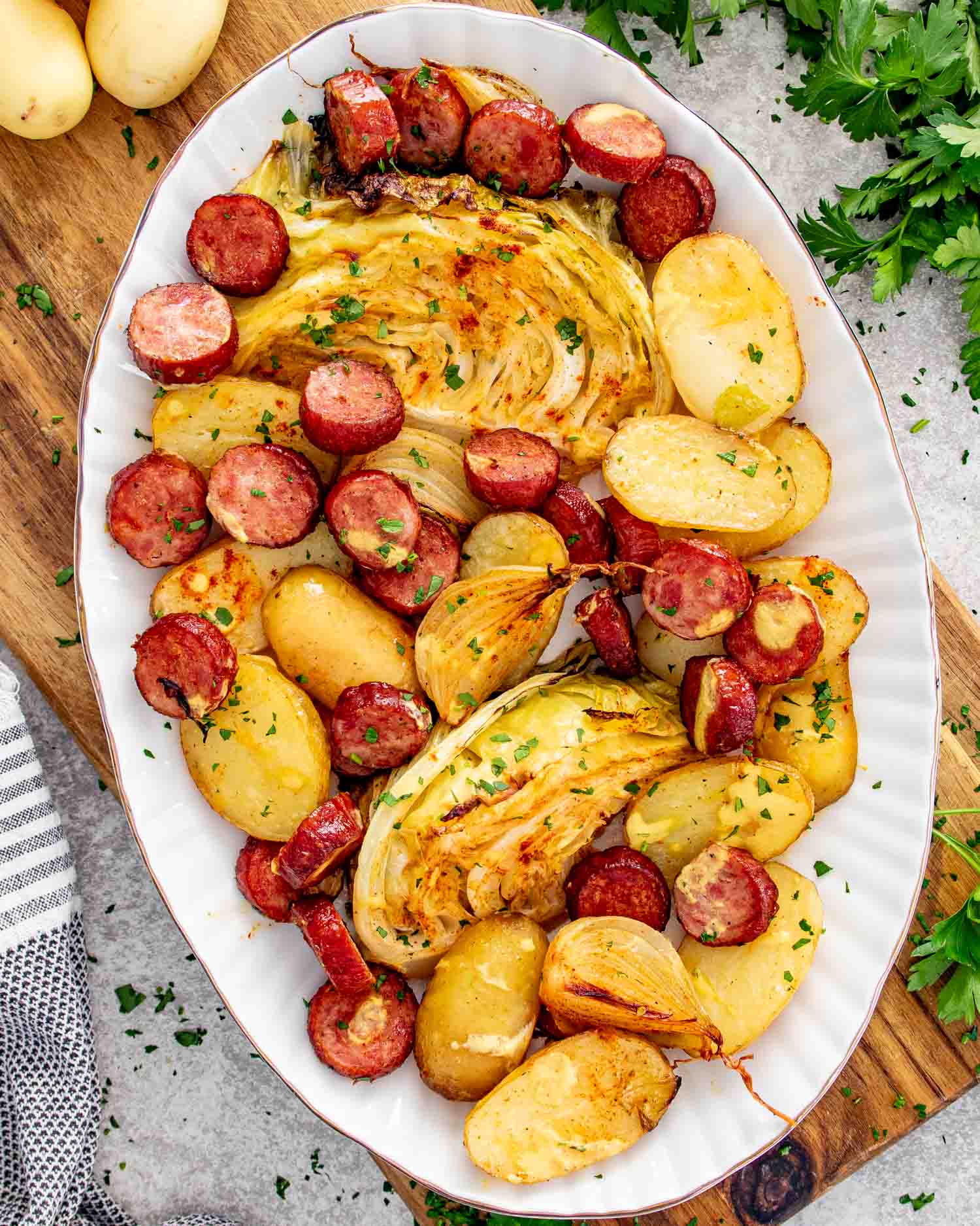 Overhead view of roasted smoked sausage, caramelized cabbage wedges, baby potatoes, and onion on a white oval serving platter garnished with fresh parsley.