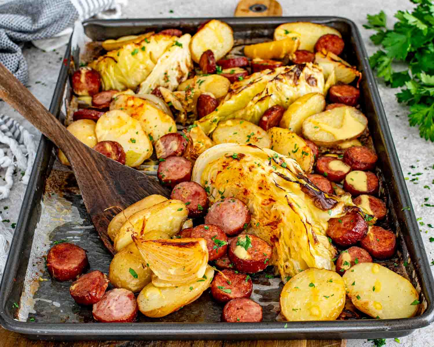 Angled view of a full sheet pan of roasted smoked sausage slices, caramelized cabbage wedges, golden baby potatoes, and onion with a wooden spatula and Dijon drizzle.