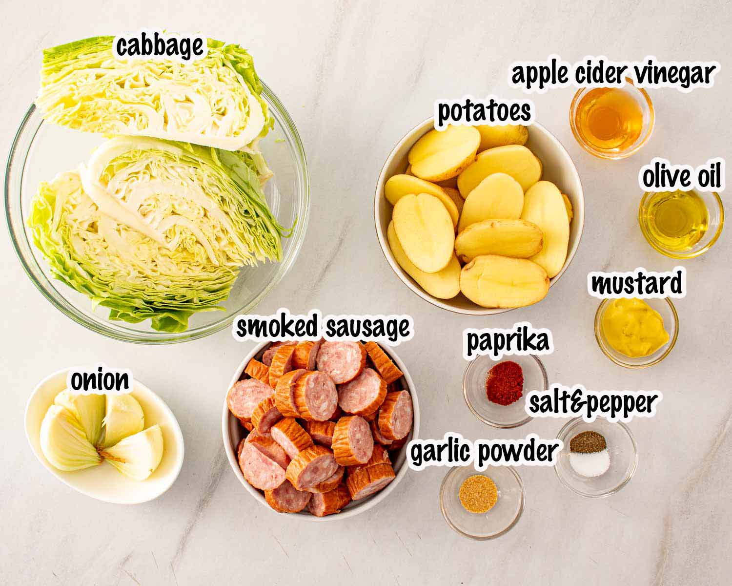 Overhead flat lay of sheet pan sausage and cabbage ingredients including smoked sausage, green cabbage wedges, baby potatoes, onion, olive oil, mustard, apple cider vinegar, paprika, garlic powder, salt, and pepper.