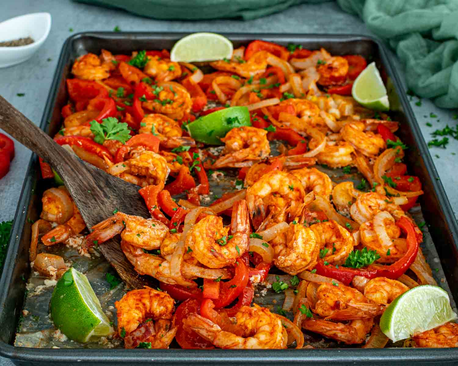 Spiced shrimp and roasted red pepper strips on a sheet pan with a wooden spatula and fresh lime wedges.