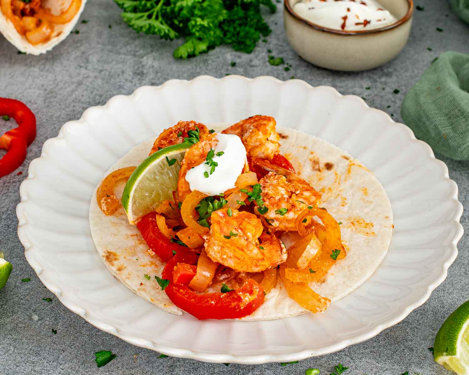 A single open shrimp fajita on a flour tortilla topped with roasted peppers, onions, a dollop of sour cream, and a lime wedge on a white scalloped plate.
