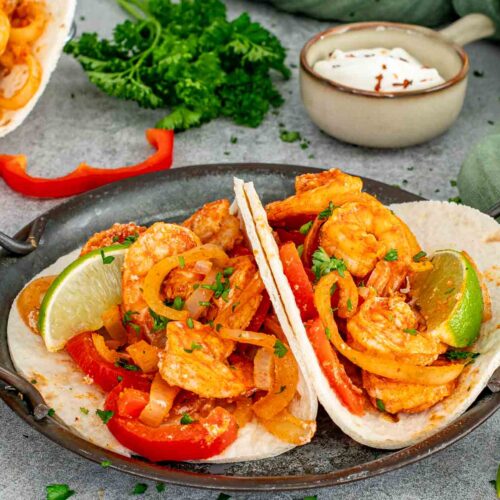 Two open shrimp fajitas loaded with red peppers and onions served in a rustic metal dish with lime wedges and sour cream in the background.