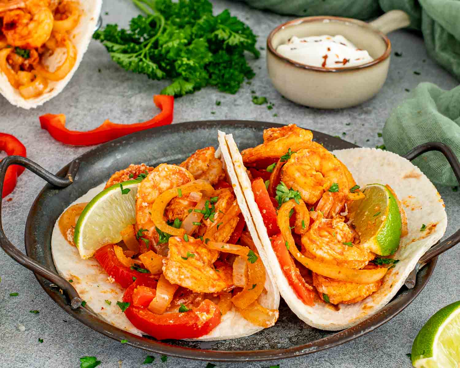 Two open shrimp fajitas loaded with red peppers and onions served in a rustic metal dish with lime wedges and sour cream in the background.