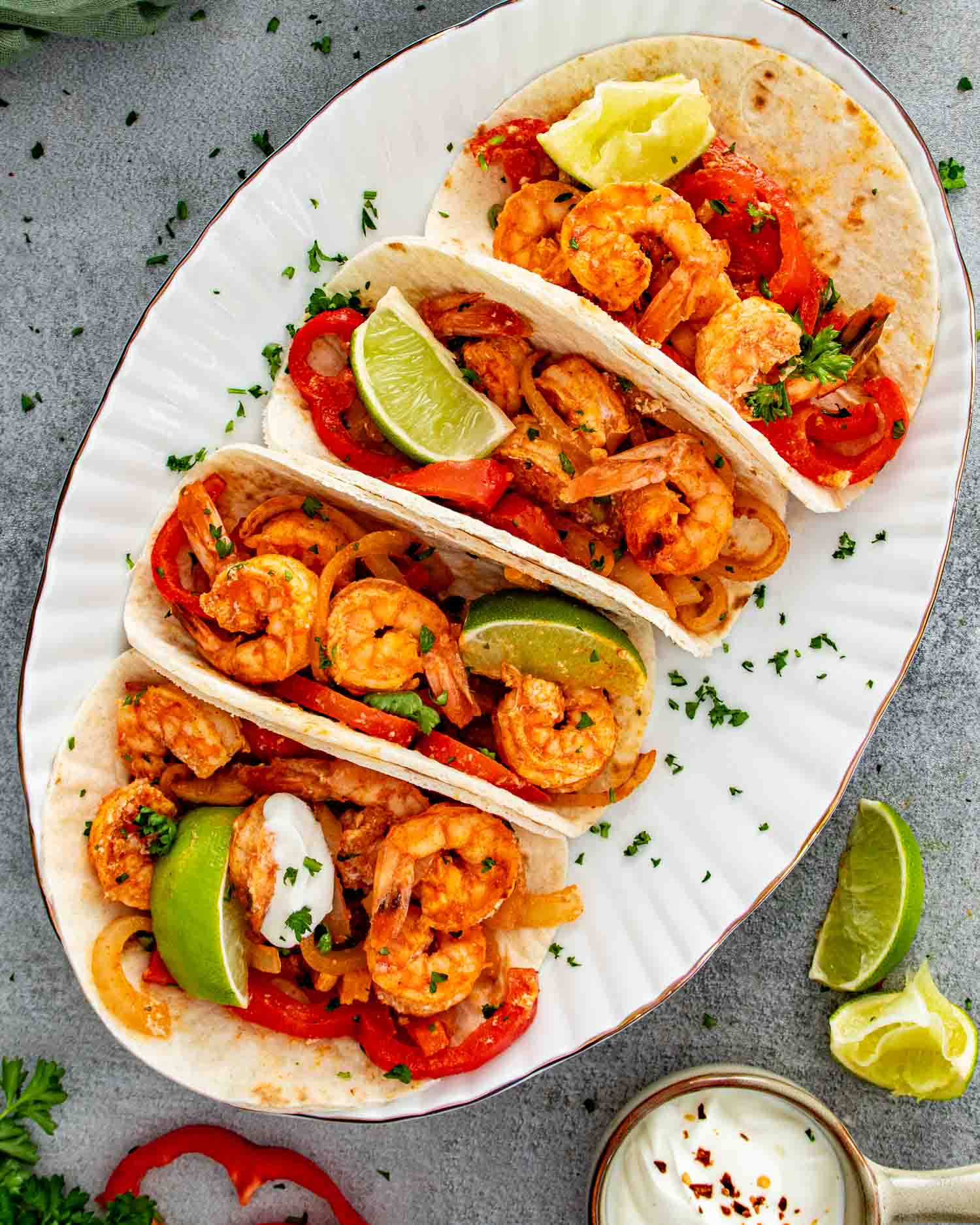 Overhead view of four shrimp fajitas on a white oval platter with roasted peppers, lime wedges, and a side of sour cream.