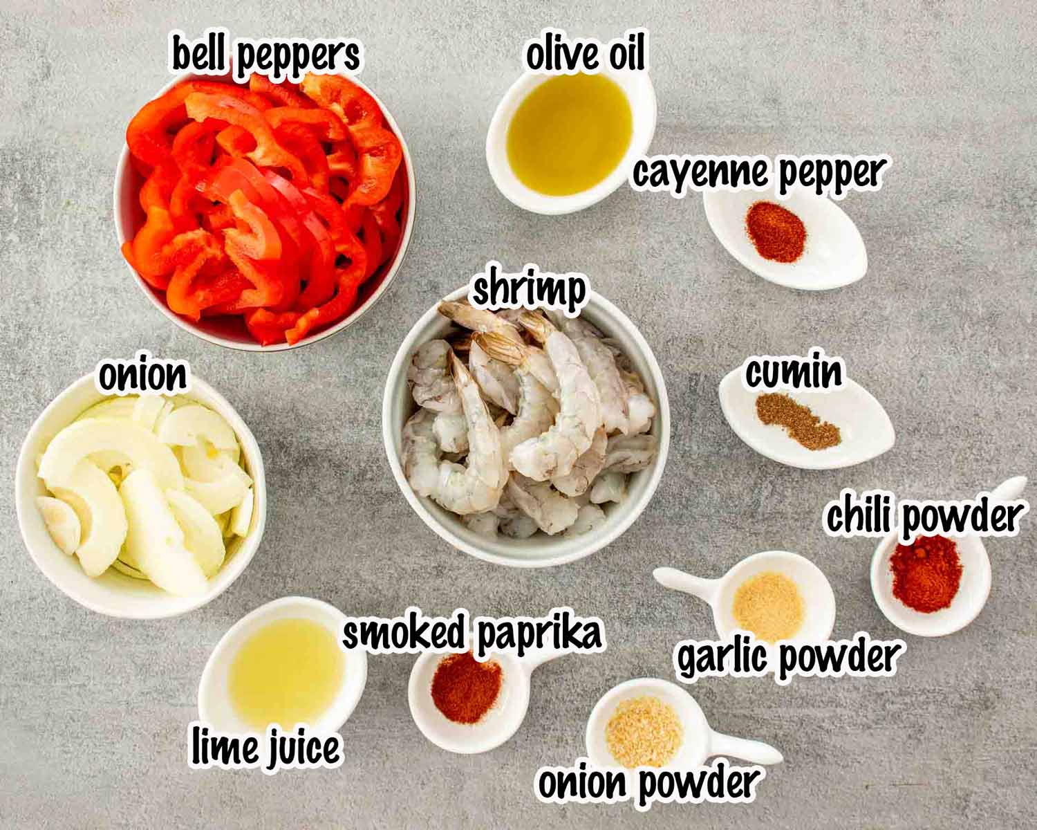 Flat lay of shrimp fajita ingredients including raw shrimp, sliced red bell peppers, onion, lime juice, olive oil, and individual spice portions labeled on a gray surface.