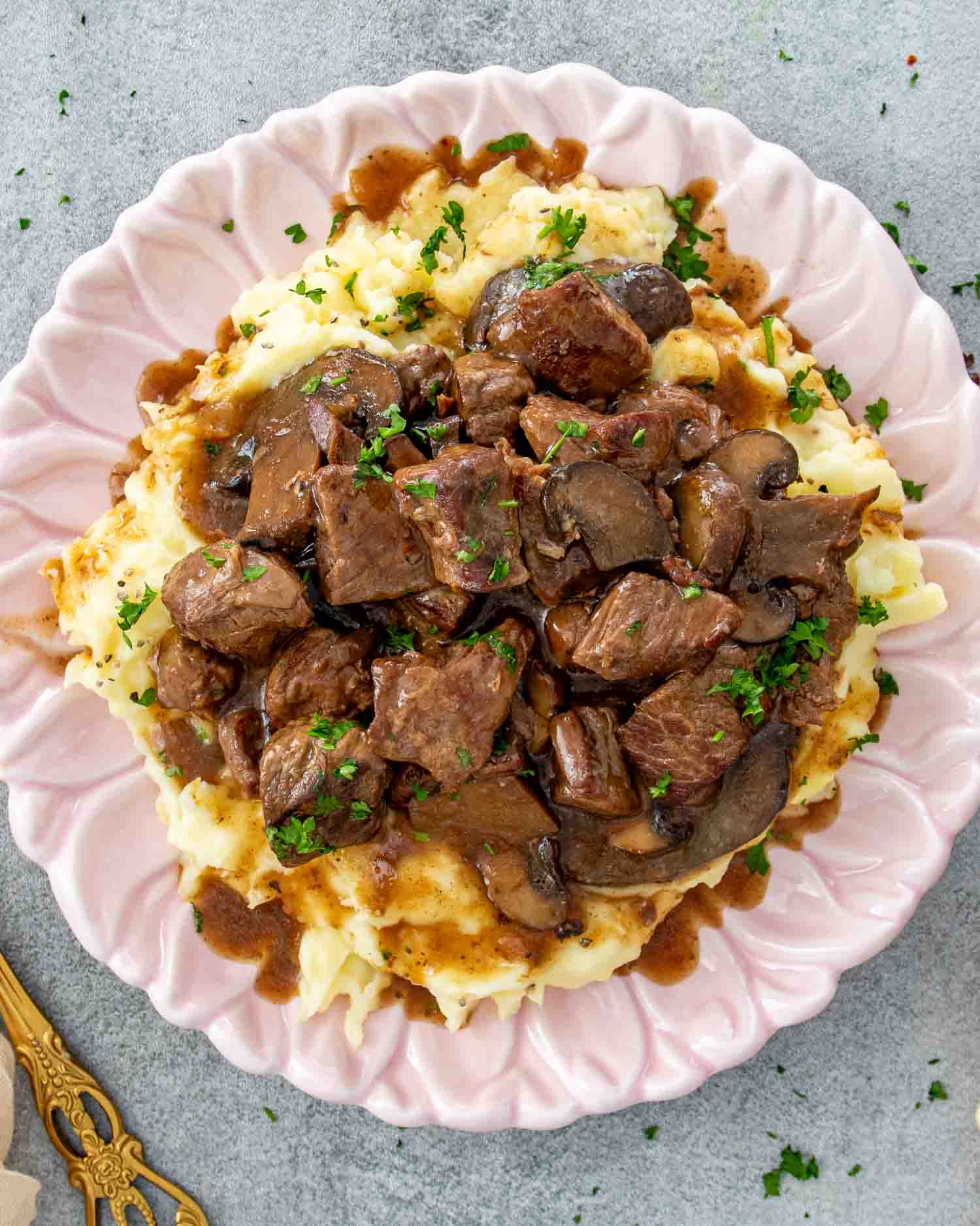 Overhead view of slow cooker beef with mushroom gravy piled over creamy mashed potatoes on a pink scalloped plate garnished with fresh parsley and a gold fork alongside.