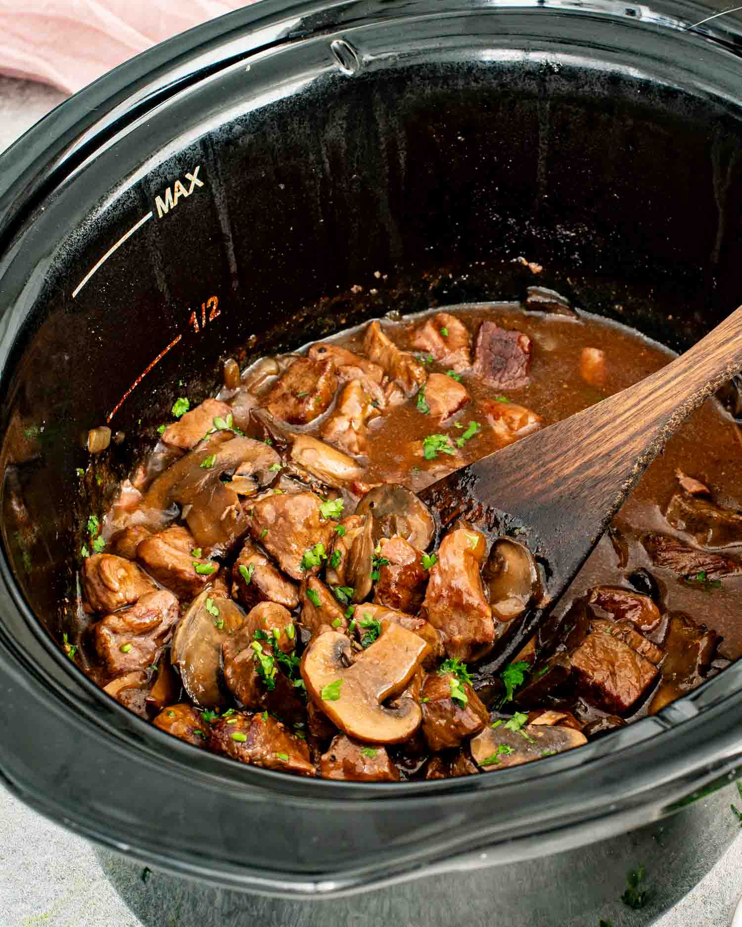 Tender beef chunks and sliced cremini mushrooms in a rich dark mushroom gravy inside a black slow cooker with a wooden spoon and fresh parsley.