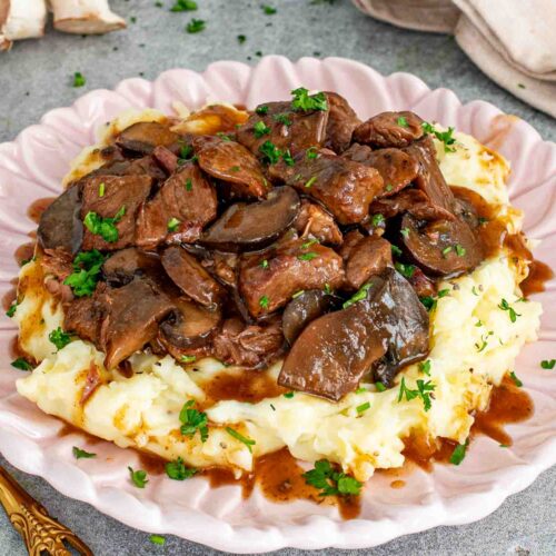 Slow cooker beef chuck and cremini mushrooms in a rich brown gravy served over mashed potatoes on a pink scalloped plate garnished with fresh parsley.