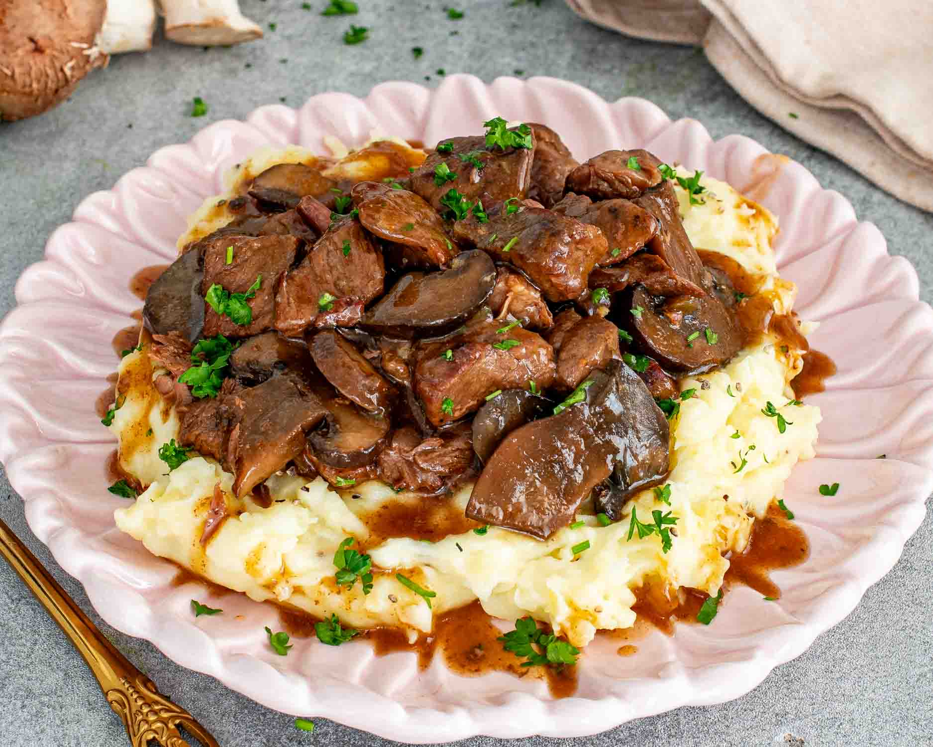 Slow cooker beef chuck and cremini mushrooms in a rich brown gravy served over mashed potatoes on a pink scalloped plate garnished with fresh parsley.