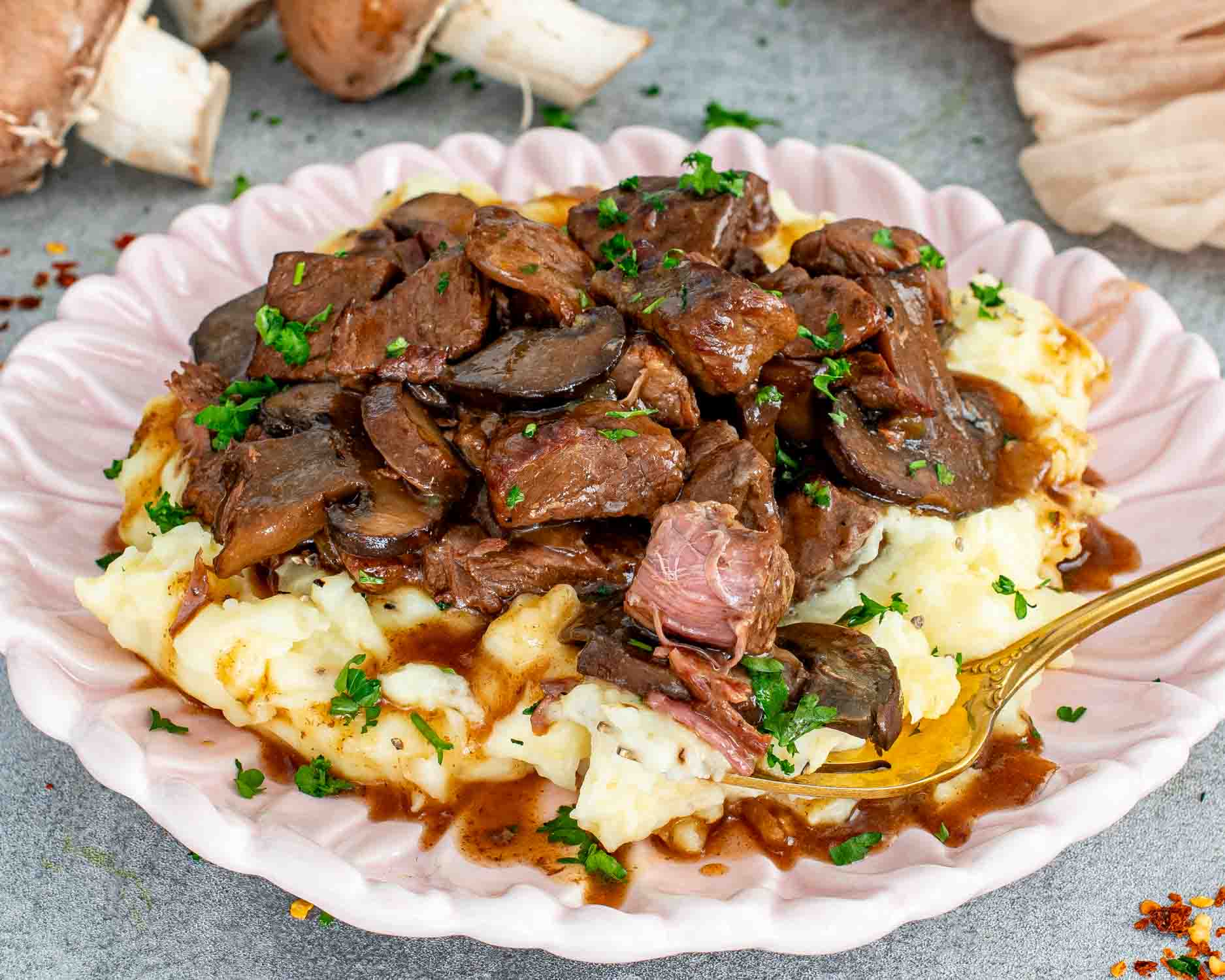 Slow cooker beef with mushroom gravy over creamy mashed potatoes on a pink scalloped plate with a gold fork lifting a tender piece of beef.