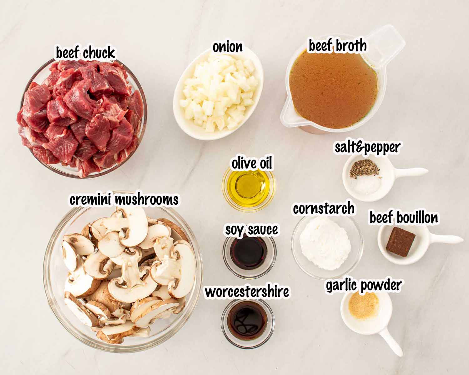 Overhead flat lay of slow cooker beef with mushroom gravy ingredients including beef chuck, cremini mushrooms, beef broth, onion, Worcestershire, soy sauce, cornstarch, beef bouillon, garlic powder, olive oil, salt, and pepper.