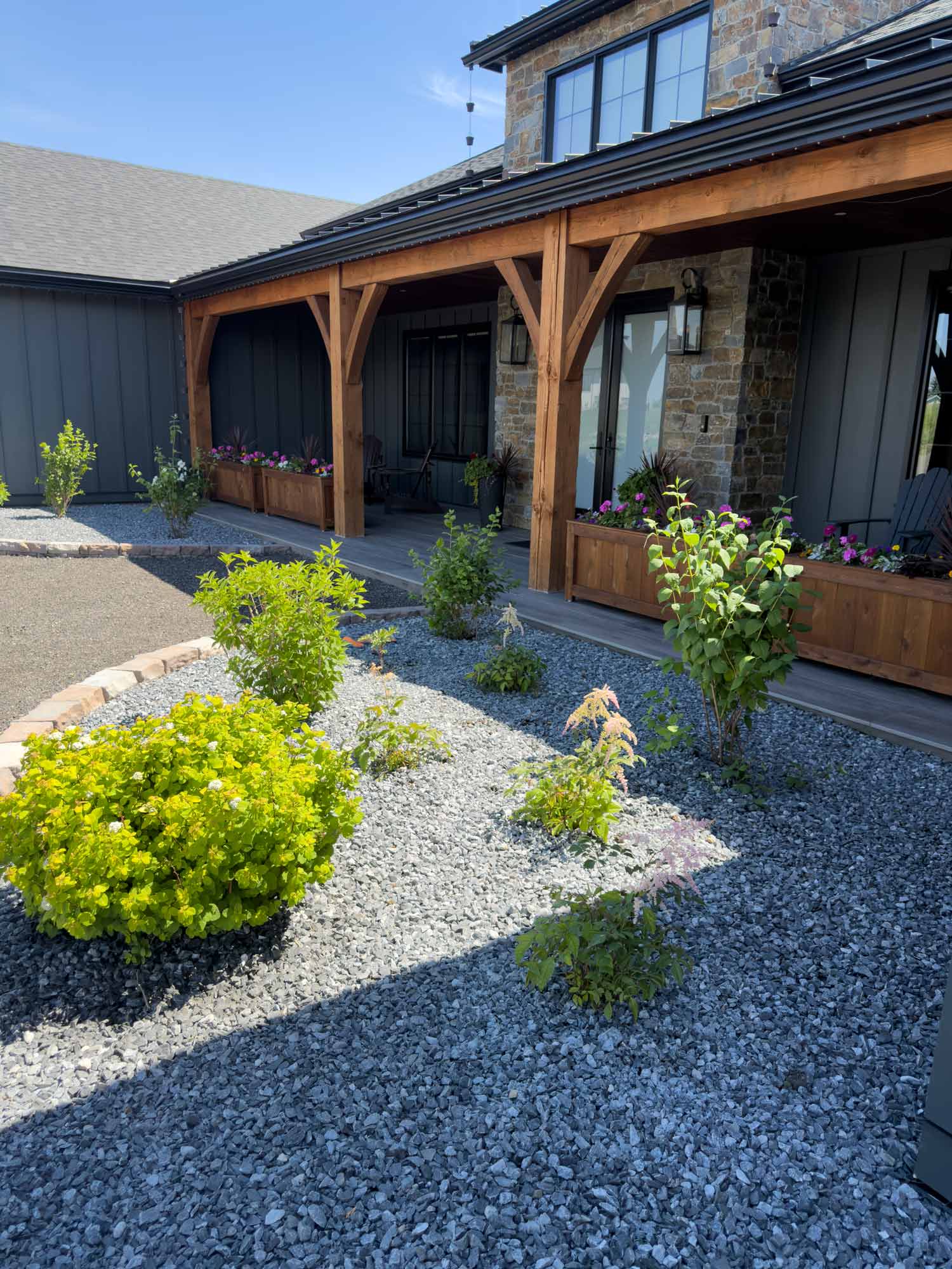 Modern farmhouse front porch with timber posts, flower boxes, fresh landscaping, shrubs, and decorative gravel surrounding welcoming front entrance.