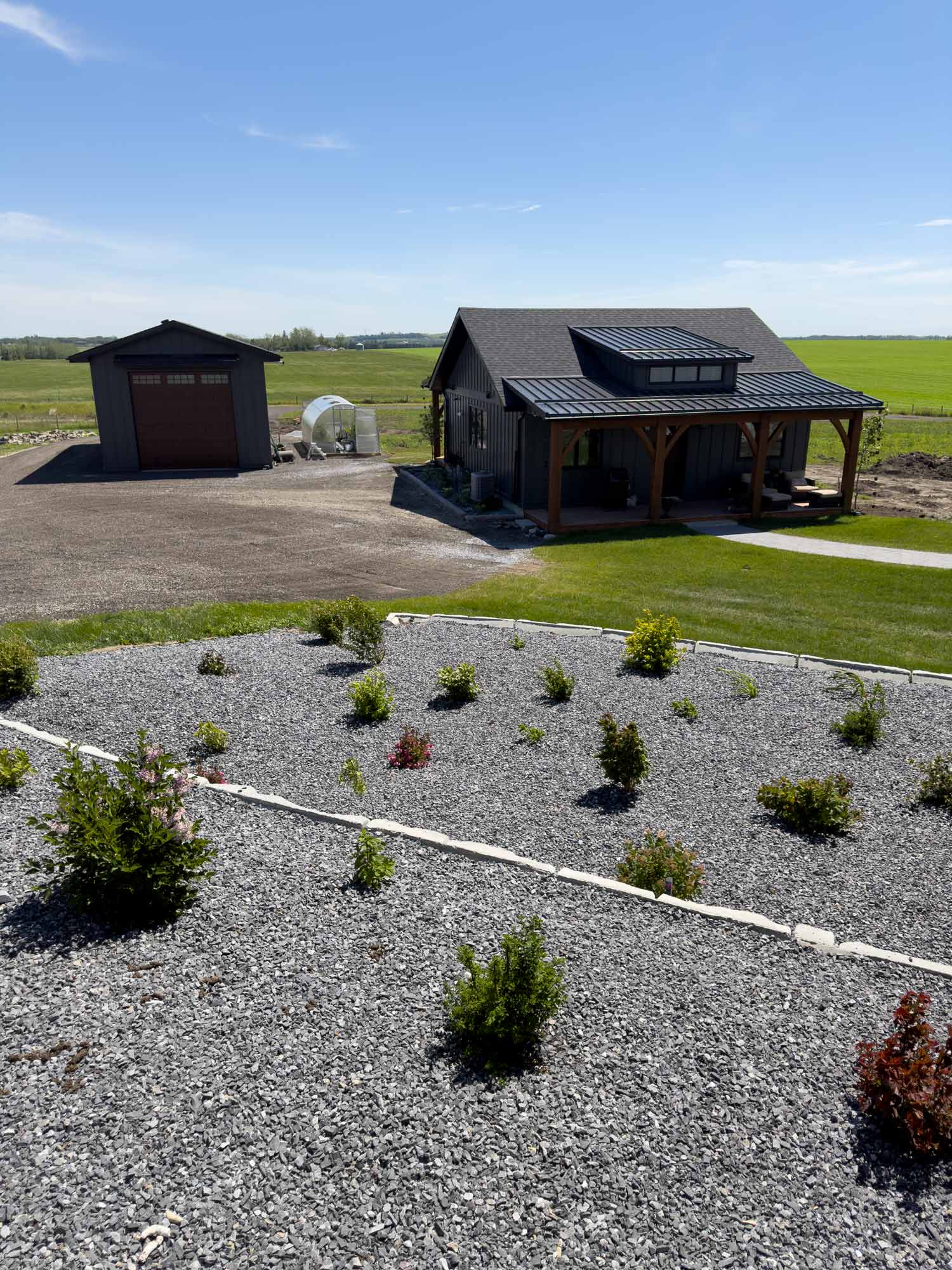 Back studio kitchen, detached garage, and greenhouse overlooking landscaped gravel beds with expansive green fields stretching into the distance.