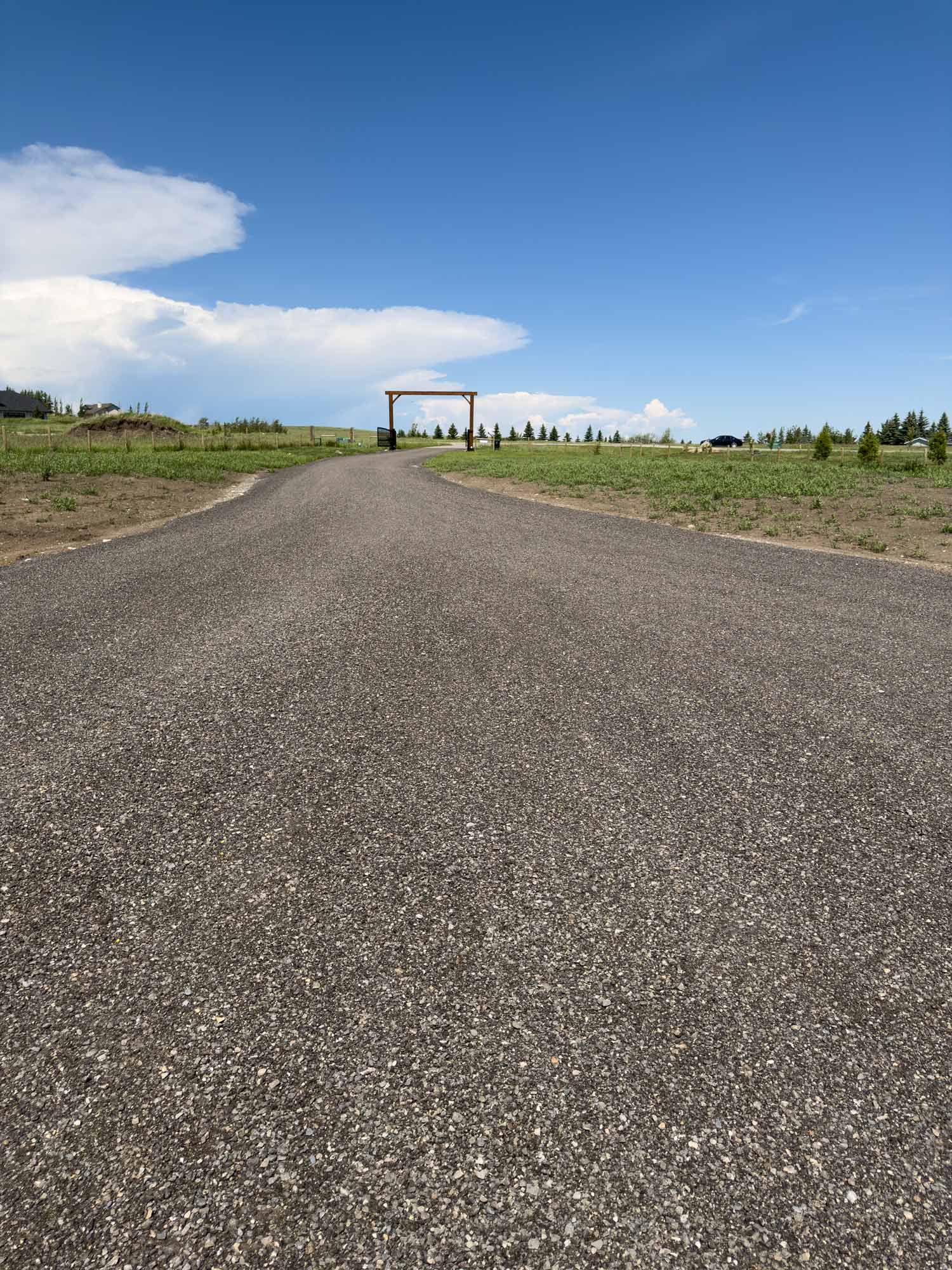 Long paved driveway leading toward rustic entrance gate with expansive prairie fields and dramatic Alberta summer clouds in the distance.