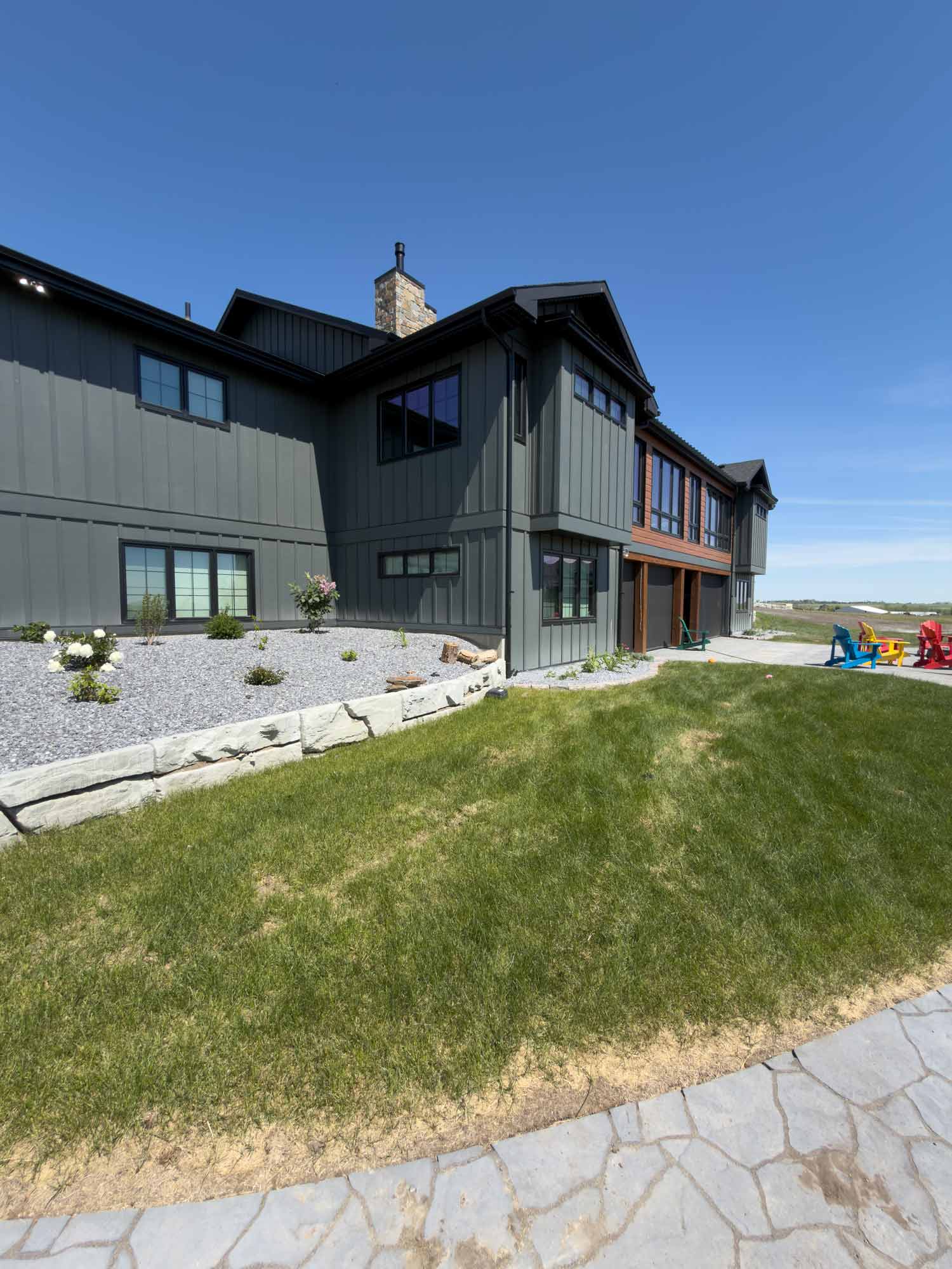 Side view of modern farmhouse featuring dark siding, stone chimney, landscaped garden beds, and expansive lawn under sunny Alberta skies.