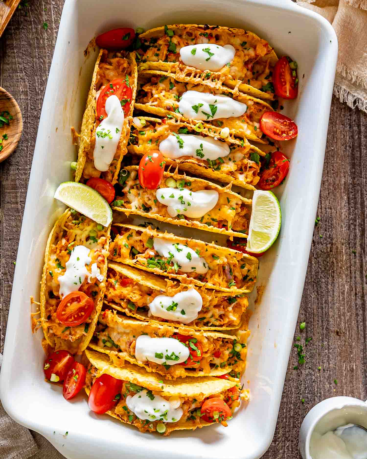 Overhead view of baked chicken tacos topped with sour cream, cherry tomatoes, herbs, and lime wedges in casserole dish.