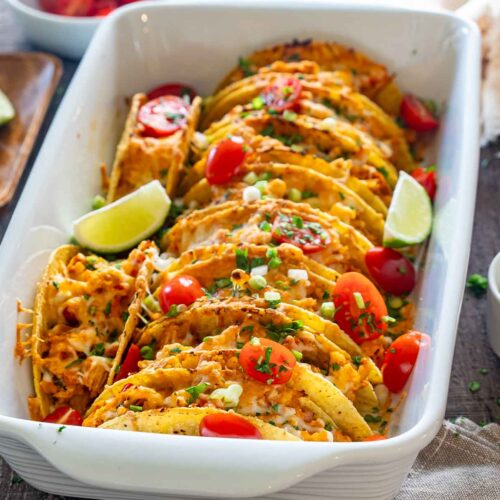 Close-up of cheesy baked chicken tacos in a white casserole dish with tomatoes, green onions, and lime wedges.
