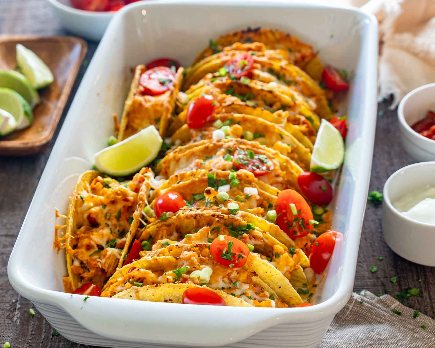 Close-up of cheesy baked chicken tacos in a white casserole dish with tomatoes, green onions, and lime wedges.