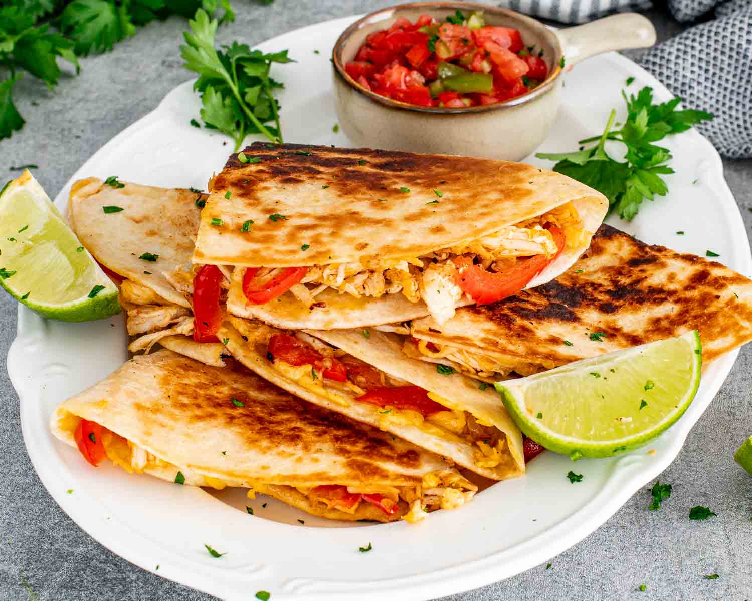Crispy chicken quesadillas folded and arranged on a white scalloped plate showing the shredded chicken and red bell pepper filling with fresh salsa and lime wedges alongside.