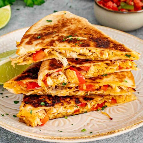 Close up of stacked crispy chicken quesadilla triangles on a ceramic plate showing the shredded chicken and red pepper filling with a bowl of salsa and lime wedges in the background.