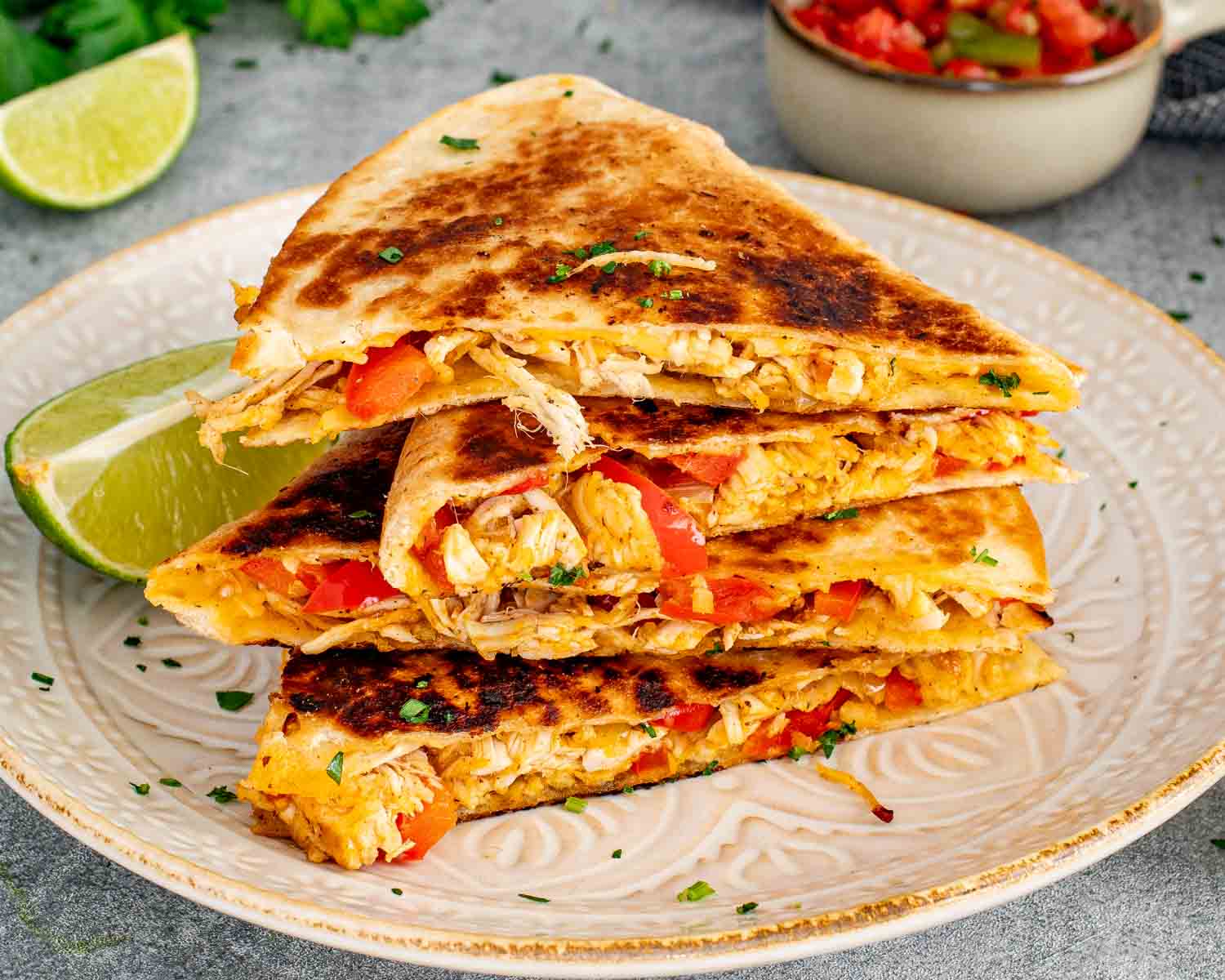 Close up of stacked crispy chicken quesadilla triangles on a ceramic plate showing the shredded chicken and red pepper filling with a bowl of salsa and lime wedges in the background.