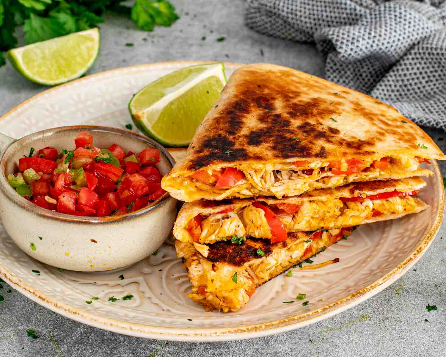 Crispy chicken quesadilla slices stacked on a ceramic plate showing the seasoned chicken and bell pepper filling served with fresh salsa and lime wedges.