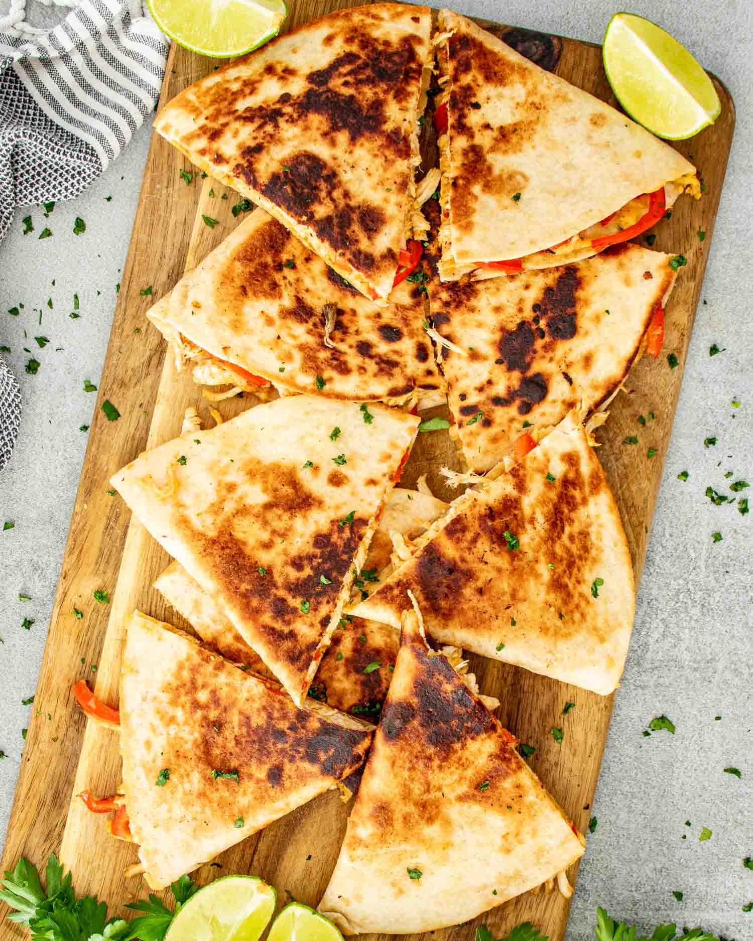 Overhead view of crispy chicken quesadillas sliced into triangles arranged on a wooden cutting board with lime wedges and fresh parsley scattered around.