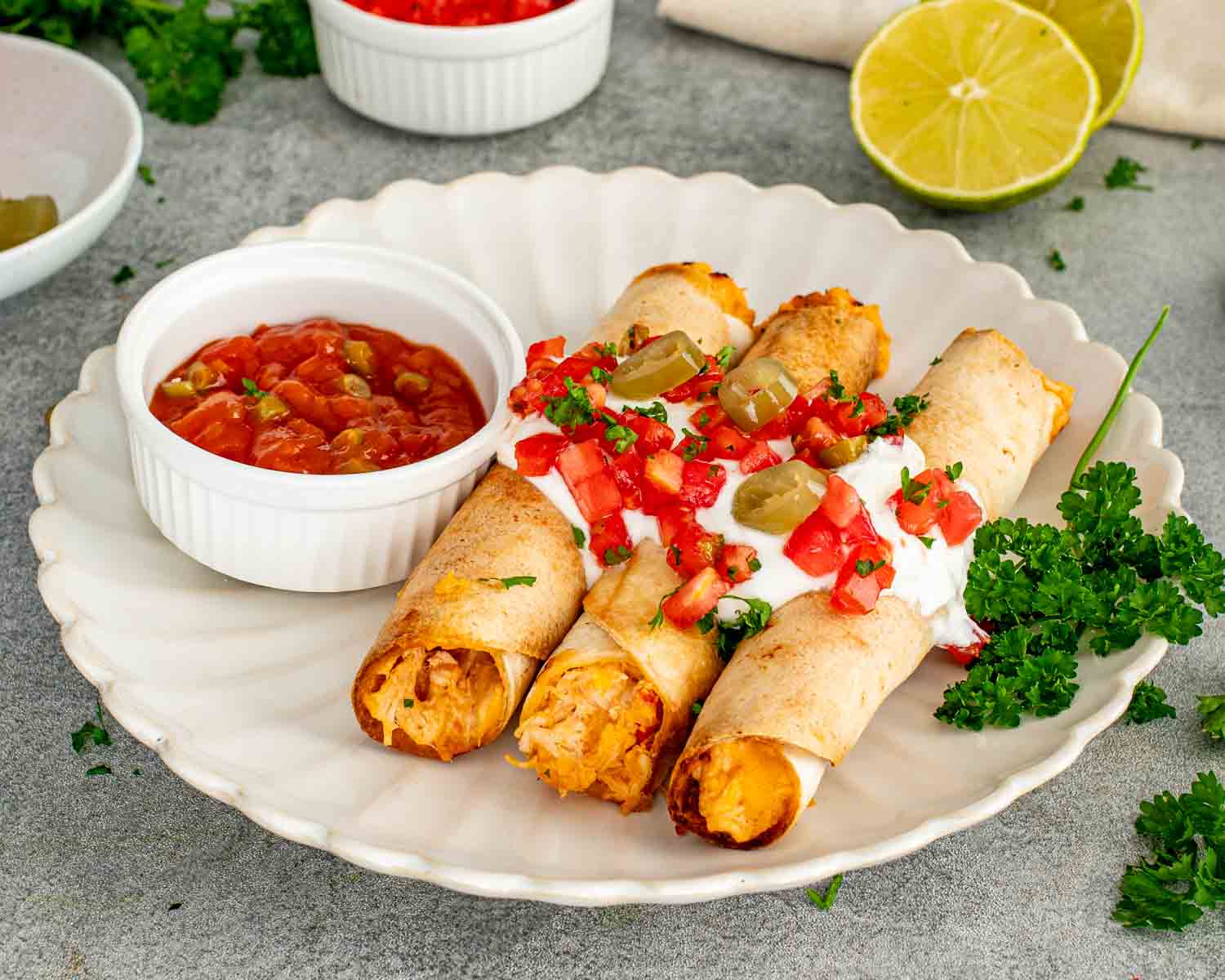 Three crispy chicken taquitos topped with sour cream, pico de gallo, jalapeños, and served alongside chunky salsa for dipping.