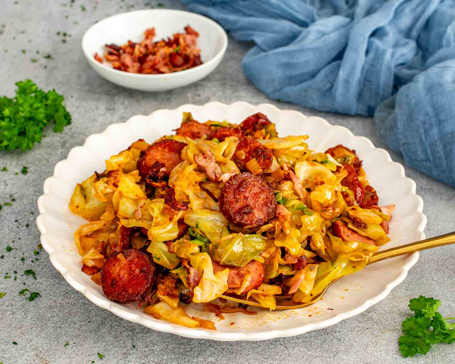 Golden fried cabbage with bacon and sausage served in a scalloped bowl with a gold serving spoon.