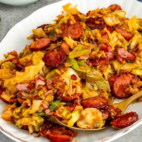 Fried cabbage with bacon and sausage served in a white bowl with a gold spoon and parsley garnish.