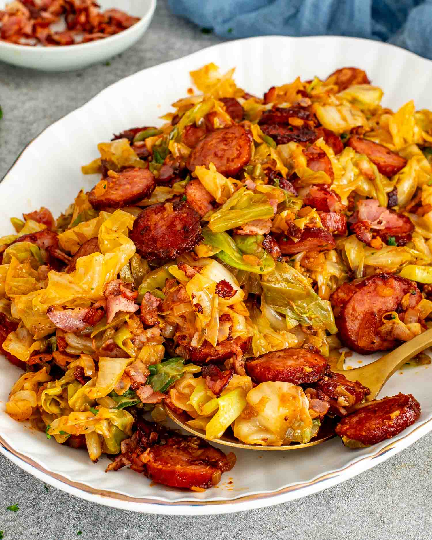 Fried cabbage with bacon and sausage served in a white bowl with a gold spoon and parsley garnish.